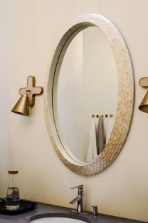 An elegant bathroom setting with a large oval mirror featuring a textured frame. On either side, there are wall-mounted, gold-toned light fixtures. Below the mirror is a modern sink with a sleek faucet. The countertop holds a glass carafe with a wooden lid. In the background, a few towels are neatly hung.