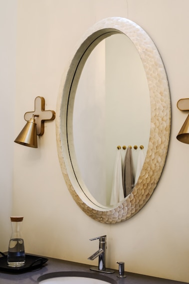 An elegant bathroom setting with a large oval mirror featuring a textured frame. On either side, there are wall-mounted, gold-toned light fixtures. Below the mirror is a modern sink with a sleek faucet. The countertop holds a glass carafe with a wooden lid. In the background, a few towels are neatly hung.