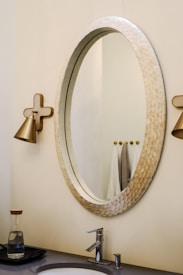 An elegant bathroom setting with a large oval mirror featuring a textured frame. On either side, there are wall-mounted, gold-toned light fixtures. Below the mirror is a modern sink with a sleek faucet. The countertop holds a glass carafe with a wooden lid. In the background, a few towels are neatly hung.