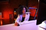 A young woman with curly hair and glasses is sitting at a desk with a computer, keyboard, and mouse. She is speaking into a microphone mounted on a stand. The background features exposed brick walls and a stop sign, with warm lighting creating an ambient atmosphere.