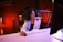 A young woman with curly hair and glasses is sitting at a desk with a computer, keyboard, and mouse. She is speaking into a microphone mounted on a stand. The background features exposed brick walls and a stop sign, with warm lighting creating an ambient atmosphere.