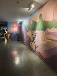 A large mural painted on an interior wall depicting various elements: on the left, a character with exaggerated features, covering their eyes with their hands in a dramatic manner, wearing a navy blue outfit with yellow details. Towards the right of the mural, there is a large bird perched on a branch. The background of the mural features elements of a green park with trees and pathways, blended with abstract architectural structures. Ceiling lights illuminate the artwork, casting shadows and highlighting the colors.