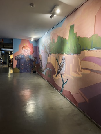 A large mural painted on an interior wall depicting various elements: on the left, a character with exaggerated features, covering their eyes with their hands in a dramatic manner, wearing a navy blue outfit with yellow details. Towards the right of the mural, there is a large bird perched on a branch. The background of the mural features elements of a green park with trees and pathways, blended with abstract architectural structures. Ceiling lights illuminate the artwork, casting shadows and highlighting the colors.