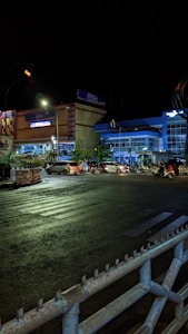 A nighttime city street scene with several cars and motorcycles waiting at a traffic light. The brightly lit buildings in the background include a bank identified by the letters BRI. The street is framed by a railing in the foreground, with zebra crossing lines visible on the road.