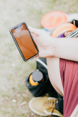 A person is holding a smartphone displaying the UDisc app. The person is wearing red shorts and has a bottle and some disc golf discs in a bag placed nearby.