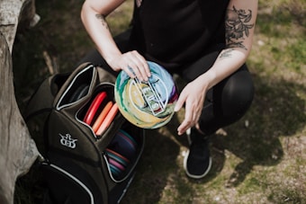 A person is squatting on the grass holding a colorful disc in their hand. They are dressed in black and have tattoos on their arms. A disc golf bag filled with various discs is open beside them on the ground.