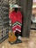 A mannequin dressed in a traditional Mexican outfit stands against a decorative background. It wears a colorful sombrero and a brightly striped poncho in shades of red, green, and black. The mannequin holds a classical Spanish guitar.