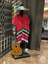 A mannequin dressed in a traditional Mexican outfit stands against a decorative background. It wears a colorful sombrero and a brightly striped poncho in shades of red, green, and black. The mannequin holds a classical Spanish guitar.