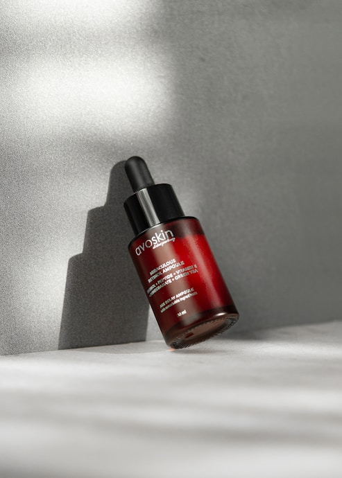 A tilted red bottle of skincare serum with a black dropper cap rests against a neutral gray background, casting a shadow on the surface. The bottle has white text labeling it as a retinol-based product.