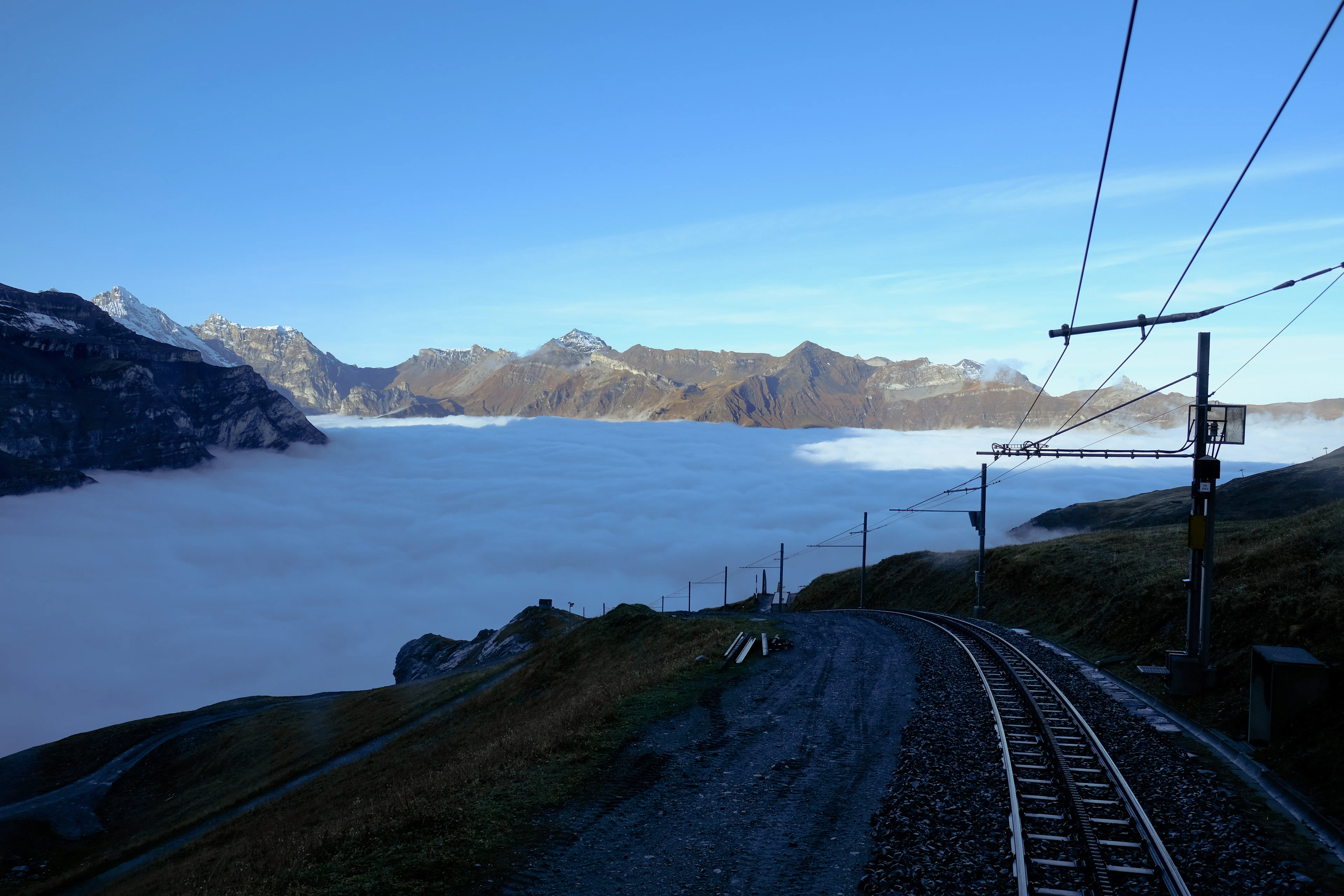 A mountain railway track curves along a ridge above a thick sea of clouds, with peaks piercing through.
