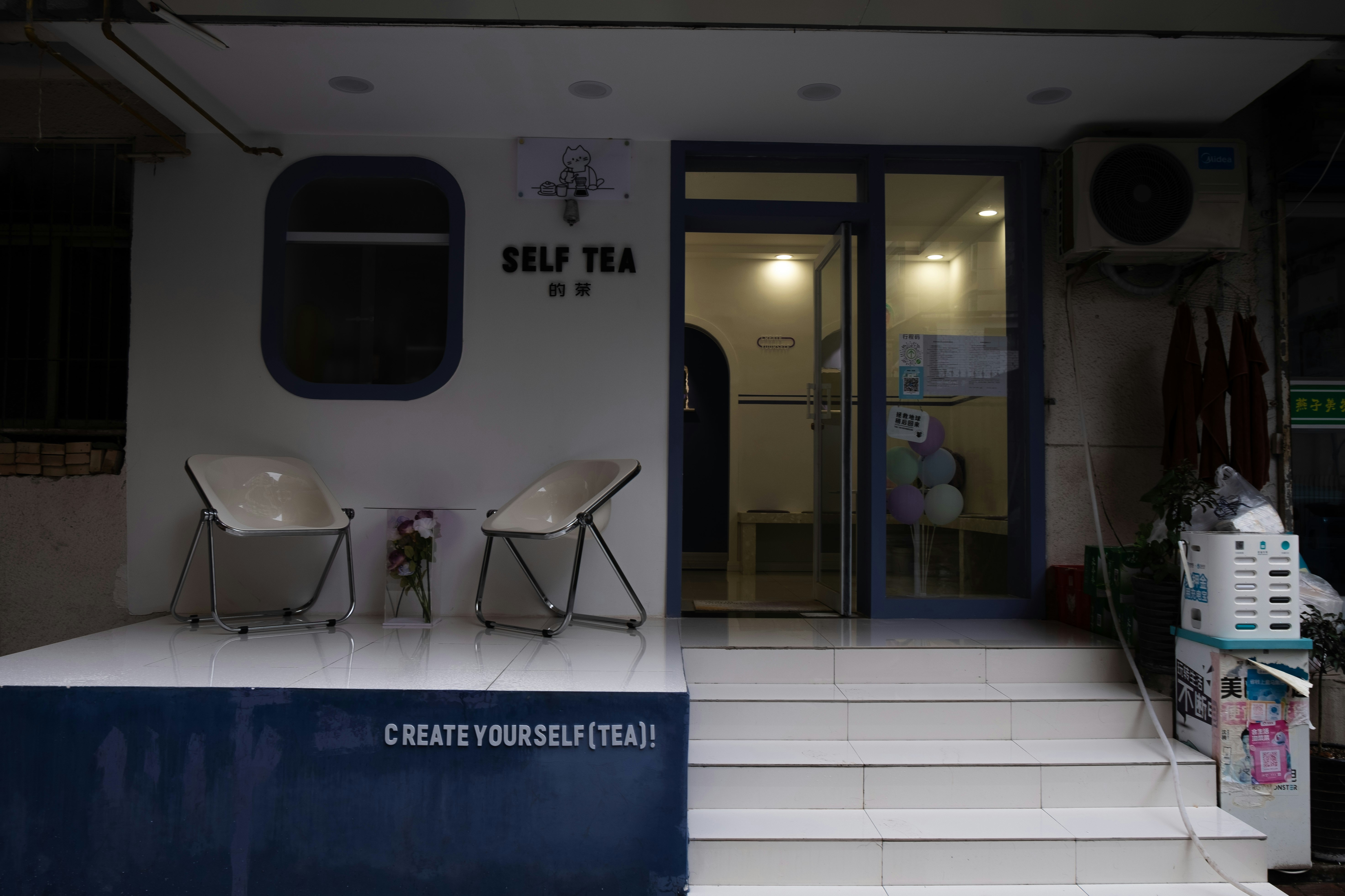 The image depicts the entrance of a tea shop named 'Self Tea'. Two modern chairs are placed on a white platform outside the shop. There is a decorative plant between the chairs. The entrance has a glass door and some colorful balloons are visible inside. A sign above the door reads 'CREATE YOURSELF (TEA)!'.