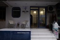 The image depicts the entrance of a tea shop named 'Self Tea'. Two modern chairs are placed on a white platform outside the shop. There is a decorative plant between the chairs. The entrance has a glass door and some colorful balloons are visible inside. A sign above the door reads 'CREATE YOURSELF (TEA)!'.