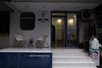 The image depicts the entrance of a tea shop named 'Self Tea'. Two modern chairs are placed on a white platform outside the shop. There is a decorative plant between the chairs. The entrance has a glass door and some colorful balloons are visible inside. A sign above the door reads 'CREATE YOURSELF (TEA)!'.