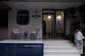 The image depicts the entrance of a tea shop named 'Self Tea'. Two modern chairs are placed on a white platform outside the shop. There is a decorative plant between the chairs. The entrance has a glass door and some colorful balloons are visible inside. A sign above the door reads 'CREATE YOURSELF (TEA)!'.