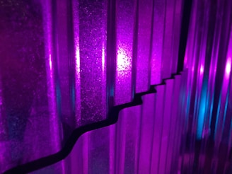 Vertical corrugated panels are illuminated with vivid purple and blue lights, creating a striking interplay of colors and shadows. The light reflects off the textures, enhancing the depth and patterns on the surface.