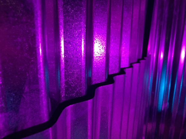 Vertical corrugated panels are illuminated with vivid purple and blue lights, creating a striking interplay of colors and shadows. The light reflects off the textures, enhancing the depth and patterns on the surface.