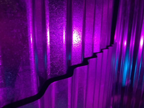 Vertical corrugated panels are illuminated with vivid purple and blue lights, creating a striking interplay of colors and shadows. The light reflects off the textures, enhancing the depth and patterns on the surface.