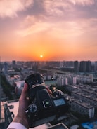 A hand is holding a DSLR camera over a cityscape during sunset. The city is filled with residential and commercial buildings extending into the horizon. The sky is tinged with shades of orange and pink as the sun sets against the skyline.