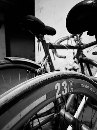 A close-up view of two bicycles with a focus on the wheel displaying the number 23C. The background is slightly out of focus, highlighting the seats and handlebars. The image is in black and white, creating a classic and timeless feel.