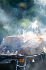 Juicy pieces of meat are being grilled over an open flame with smoke rising up, indicating they are being cooked thoroughly. The background is slightly blurred, creating a focus on the grilling process.