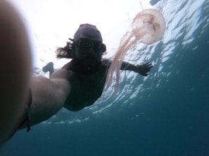 A person is snorkeling underwater, extending an arm forward with a visible wristband. A jellyfish floats nearby, its tentacles trailing through the water. The water appears clear with light reflections on the surface.