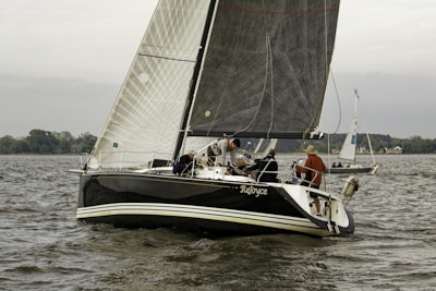 A sailboat with the name 'ReJoyce' is navigating through a body of water with several people onboard. The sailing vessel has dark-colored hull and sails, and others are also sailing in the background. The scene is set near the shoreline, with greenery and some structures visible in the distance. The overall atmosphere seems calm with a slightly overcast sky.