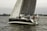 A sailboat with the name 'ReJoyce' is navigating through a body of water with several people onboard. The sailing vessel has dark-colored hull and sails, and others are also sailing in the background. The scene is set near the shoreline, with greenery and some structures visible in the distance. The overall atmosphere seems calm with a slightly overcast sky.