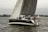 A sailboat with the name 'ReJoyce' is navigating through a body of water with several people onboard. The sailing vessel has dark-colored hull and sails, and others are also sailing in the background. The scene is set near the shoreline, with greenery and some structures visible in the distance. The overall atmosphere seems calm with a slightly overcast sky.