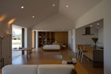 A spacious and modern open-plan living room with natural lighting, featuring a wooden floor, a white ceiling with recessed lights, and a large sliding door that opens to an outdoor area. The room includes a bed against a built-in shelving unit on one side and a minimalist kitchen with wooden elements and a sleek island counter on the other. A comfortable sofa is positioned near a small coffee table.