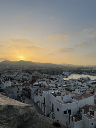 Sunset over a coastal cityscape comprising white buildings with terracotta roofs, a marina with boats, and surrounding hills. The sky transitions from yellow to blue as the sun sets behind the mountains.