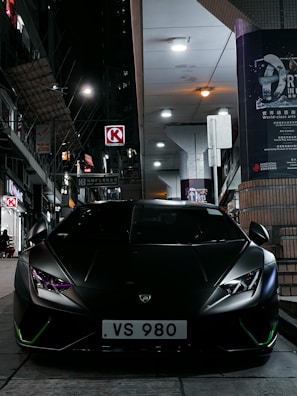 A sleek, black Lamborghini is parked on a city street at night, with modern buildings and illuminated signs in the background, creating an urban atmosphere. The headlights of the car are on, producing a sharp, focused look.