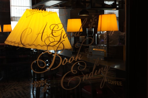 A warm-lit cafe interior with decorative script lettering on a glass window that reads 'A Good Book Ending.' Several yellow lampshades hang above seating areas, casting a cozy ambiance.