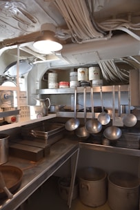 A kitchen area with various cooking utensils and equipment. Shelves hold cans labeled with ingredients like eggs, beets, and camel. Large ladles hang over metal countertops, and pots are stored below. Overhead, wiring is visible alongside a light fixture.