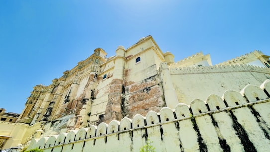 A majestic palace with intricate architectural details, featuring a blend of white and sandstone colors. The structure is adorned with decorative elements and has a high facade that stretches upwards into a clear blue sky.