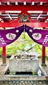 A traditional Japanese purification fountain located under a vibrant red and purple structure. The roof is decorated with ornate patterns and an emblem featuring an eagle, while purple drapes with white symbols hang underneath. Bamboo ladles rest on top of the stone basin, and a serene pond with lush greenery is visible in the background.