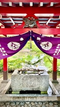 A traditional Japanese purification fountain located under a vibrant red and purple structure. The roof is decorated with ornate patterns and an emblem featuring an eagle, while purple drapes with white symbols hang underneath. Bamboo ladles rest on top of the stone basin, and a serene pond with lush greenery is visible in the background.