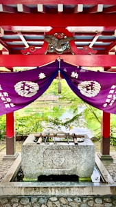 A traditional Japanese purification fountain located under a vibrant red and purple structure. The roof is decorated with ornate patterns and an emblem featuring an eagle, while purple drapes with white symbols hang underneath. Bamboo ladles rest on top of the stone basin, and a serene pond with lush greenery is visible in the background.