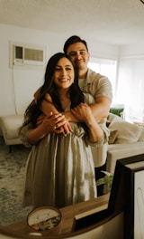A couple is embracing each other warmly in a cozy, softly lit living room with a beige couch and a patterned rug. Framed pictures and a decorative candle are placed on the wooden table in the foreground. The room has a modern and comfortable ambiance with natural light streaming in through the windows.