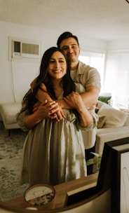A couple is embracing each other warmly in a cozy, softly lit living room with a beige couch and a patterned rug. Framed pictures and a decorative candle are placed on the wooden table in the foreground. The room has a modern and comfortable ambiance with natural light streaming in through the windows.
