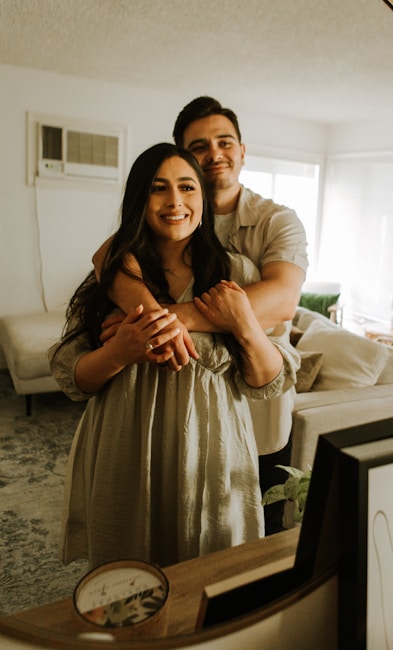 A couple is embracing each other warmly in a cozy, softly lit living room with a beige couch and a patterned rug. Framed pictures and a decorative candle are placed on the wooden table in the foreground. The room has a modern and comfortable ambiance with natural light streaming in through the windows.