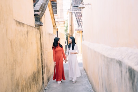 Two women in traditional attire walk hand in hand down a narrow alley with yellow walls. They appear to be engaged in a friendly conversation, enjoying a sunny day.