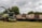 A large agricultural machine, likely a forage harvester, is parked on a grassy field. It is equipped with a header suitable for cutting crops. Behind the harvester, several large trailers are lined up, which appear to be used for transporting harvested materials. The background consists of dense green trees under an overcast sky.