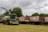 A large agricultural machine, likely a forage harvester, is parked on a grassy field. It is equipped with a header suitable for cutting crops. Behind the harvester, several large trailers are lined up, which appear to be used for transporting harvested materials. The background consists of dense green trees under an overcast sky.