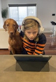 A child with headphones is focused on a tablet while a brown dog sits closely beside them. They are indoors, with a couch, plant, and barstools visible in the background.