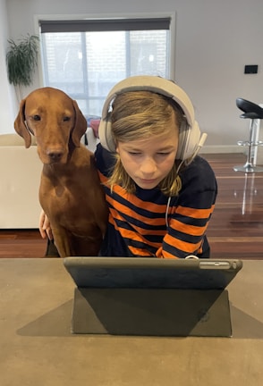 A child with headphones is focused on a tablet while a brown dog sits closely beside them. They are indoors, with a couch, plant, and barstools visible in the background.
