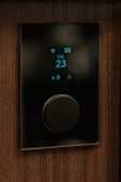 A sleek, modern thermostat with a digital display showing a temperature reading of 23 degrees. It is mounted on a wooden wall. The display includes icons for WiFi, humidity, and heating, with a large central dial for adjusting settings.