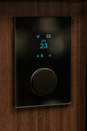 A sleek, modern thermostat with a digital display showing a temperature reading of 23 degrees. It is mounted on a wooden wall. The display includes icons for WiFi, humidity, and heating, with a large central dial for adjusting settings.