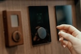 A close-up view of a person's hand adjusting a digital thermostat on a wooden wall. The thermostat has a circular knob and a small display showing the temperature. Next to it, there is a smaller device with a teardrop-shaped button.