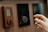 A close-up view of a person's hand adjusting a digital thermostat on a wooden wall. The thermostat has a circular knob and a small display showing the temperature. Next to it, there is a smaller device with a teardrop-shaped button.