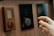 A close-up view of a person's hand adjusting a digital thermostat on a wooden wall. The thermostat has a circular knob and a small display showing the temperature. Next to it, there is a smaller device with a teardrop-shaped button.